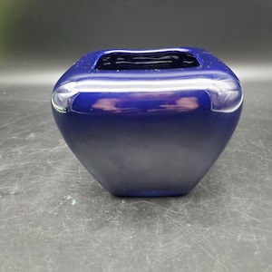 May include: A deep cobalt blue ceramic vase with a smooth, glossy finish. The vessel features a square-shaped opening with rounded corners and a tapered body that narrows toward the base, creating a sleek, modern silhouette with reflective surfaces.