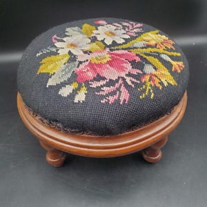 May include: A round footstool with a dark brown wooden base and four short legs. The top is upholstered in black fabric featuring a needlepoint floral design in shades of pink, white, yellow, and green. The cushion has a decorative trim edge.