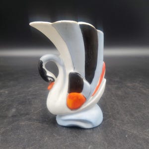 Puede incluir: Una jarra de cerámica con forma de cisne que presenta un cuerpo blanco con detalles en azul claro, negro y naranja brillante. El cuello del cisne forma el asa, mientras que sus alas y cola crean el borde acampanado y el cuerpo del recipiente.