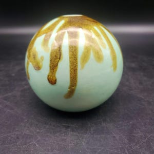 May include: A spherical ceramic vase with a smooth, light mint green glaze. The top section features a decorative brown drip pattern that cascades downward in irregular, vertical streaks, creating a contrast against the pale base color.
