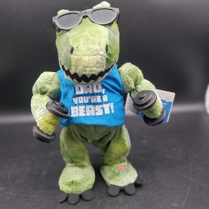 Puede incluir: Un juguete de peluche verde de un Tyrannosaurus Rex con gafas de sol y una camiseta sin mangas azul con el texto "DAD, YOU'RE A BEAST!". El juguete sostiene dos pequeñas pesas negras. Una etiqueta está adjunta.