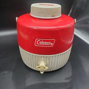 Könnte beinhalten: Eine rote und weiße Coleman-Kühlbox im Vintage-Stil. Die Kühlbox hat einen roten oberen Teil mit dem Coleman-Logo und einen weißen unteren Teil mit einem Wasserhahn. Der Deckel ist weiß. Die Kühlbox ist für das Kalthalten von Getränken konzipiert.