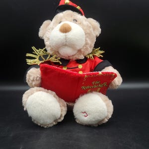 May include: A plush teddy bear wearing a red and black military-style jacket with gold epaulets and a matching cap. The bear holds an open red book with the text The Nutcracker printed on the cover. The bear has light brown fur and white paws.