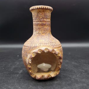 May include: A brown, textured clay candle holder with a rustic, weathered finish. The vessel features a narrow neck, a rounded body, and a large, scalloped-edge opening at the front that contains a small, round, white tea light candle inside.