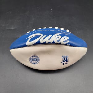 May include: A blue and white football with the word "Duke" in white script. The football has white stitching and a white lower section. Logos are printed on the white section.