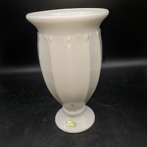 May include: A white milk glass vase with a flared rim and a vertical paneled body. The vase features a pedestal base and a small green rectangular sticker on the lower section with the text "Hagen-Renaker" printed in black lettering.