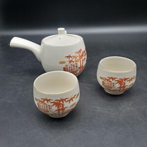 May include: A white ceramic teapot with a side handle and two matching small cups. The set features a decorative pattern of orange bamboo leaves and stalks, along with black Japanese calligraphy text printed on the surface of each piece.