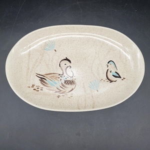 Puede incluir: Una bandeja ovalada de cerámica con un acabado en color crema moteado. El centro presenta una ilustración pintada a mano de un pato grande y un pájaro más pequeño, con detalles en marrón y azul claro, rodeados de pequeños motivos florales.