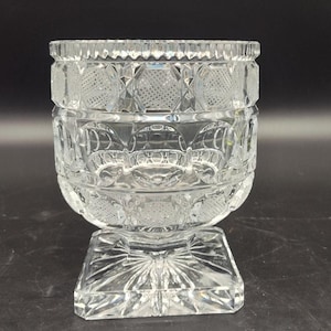 May include: A clear, cut-glass bowl with a square base and a rounded body. The bowl features intricate geometric patterns and a textured rim. The design is symmetrical and the glass is transparent, allowing light to pass through.