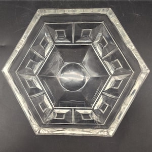 May include: A clear glass hexagonal bowl with geometric patterns. The bowl features a central circular indentation and is surrounded by diamond and square shapes. The bowl is made of clear glass and is suitable for serving or display purposes.