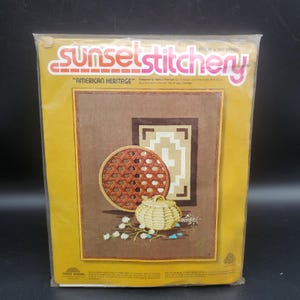 May include: A Sunset Stitchery craft kit in a clear plastic package. The kit features a brown and tan stitched design of a basket, a woven plate, and a geometric pattern. The text "Sunset Stitchery" and "American Heritage" are visible on the package.