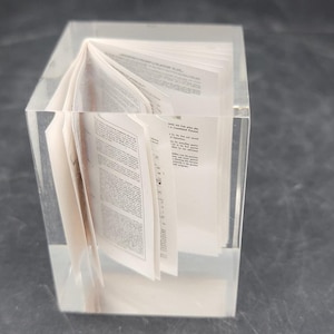 May include: A transparent acrylic block containing a small, open book. The book's pages are visible, displaying printed text. The block is clear, offering a view of the book's details. The book is positioned centrally within the block.