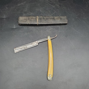 May include: A vintage straight razor with a cream-coloured handle and a silver blade. The razor is next to a black leather case. The blade is slightly curved at the end. The razor is open and ready for use.