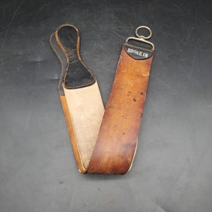 May include: A vintage leather strop with a black handle and a metal ring. The strop is brown with visible wear and the text "BROKE IN" is printed on the top. The other side is a lighter color, possibly canvas.