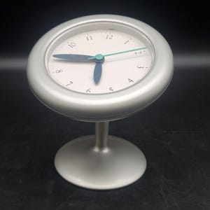 Peut inclure: Une horloge de bureau analogique argentée avec un cadran circulaire, un fond rose clair et des chiffres noirs. L'horloge présente deux aiguilles bleu foncé et une fine aiguille vert clair, montée sur un pied avec une base circulaire.