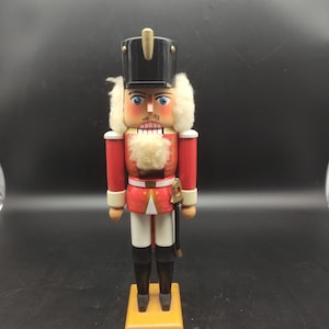 Peut inclure: Figurine de casse-noisette en bois, vêtu d'un uniforme rouge et blanc, coiffé d'un chapeau noir et orné d'une barbe blanche. Il porte une épée dorée et repose sur un socle en bois. Décoration de Noël classique.