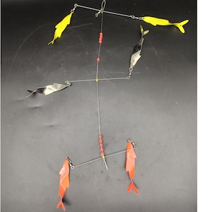 Puede incluir: Señuelo de pesca con una estructura central de alambre y múltiples brazos. Cuenta con componentes en forma de pez amarillos y naranjas, hojas reflectantes plateadas y cuentas rojas. Diseñado para atraer peces.