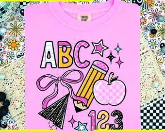 May include: A pink t-shirt with a school-themed graphic. The design includes the letters "ABC", the numbers "123", a pencil, an apple, and stars. The text "DTF TRANSFER ONLY" and "Pineapple Transfers" are also visible.