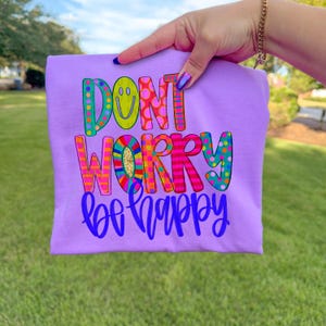 May include: A light purple t-shirt with the words "DON'T WORRY be happy" in colorful, playful lettering. The design includes a smiley face and various patterns. The shirt is held up against a green grassy background.
