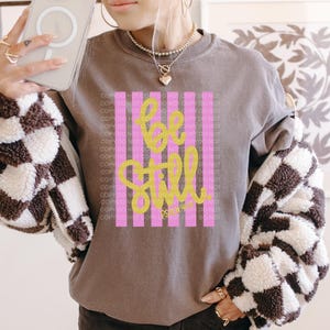 May include: A brown crew neck sweatshirt with the words "be still" in yellow script. The text is superimposed on a pink and white striped background. The person is wearing a black and white checkered jacket.