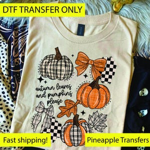 May include: Cream-colored t-shirt featuring a fall-themed graphic. The design includes orange pumpkins, plaid bows, and the text "autumn leaves and pumpkins please." The shirt is a DTF transfer.