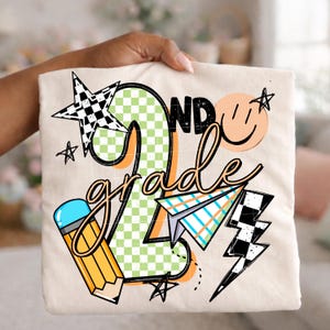 May include: A cream-colored t-shirt featuring a graphic with the text 2nd grade. The design includes a large green checkered number two, a yellow pencil, a paper airplane, a black and white checkered star, a lightning bolt, and a smiley face.