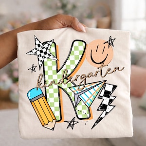 May include: A cream-colored t-shirt featuring a large green and white checkered letter K. The design includes a yellow pencil, a paper airplane, a black and white checkered star, a lightning bolt, and a peach-colored smiley face with the word kindergarten.
