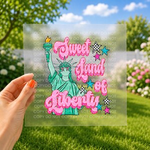 May include: A clear decal featuring a green Statue of Liberty holding a torch, with pink text reading Sweet Land of Liberty. The design includes pink and blue stars, and black and white checkered stars, all set against a transparent background.