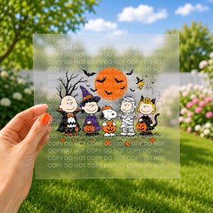 May include: A square, semi-transparent transfer sheet featuring a Peanuts Halloween illustration. Characters include Charlie Brown as a vampire, Lucy as a witch, Snoopy as a pumpkin, Linus as a mummy, and Sally as a black cat, all under an orange moon.