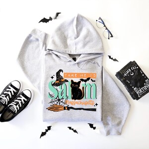 May include: A light gray hooded sweatshirt featuring a graphic print that reads Take Me To Salem Massachusetts in teal and orange text. The design includes a black witch hat, a black cat, a spider web, and a broomstick with orange bristles.