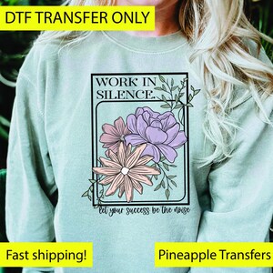 May include: A light green sweatshirt featuring a floral design with the text "WORK IN SILENCE" and "let your success be the noise". The floral design includes pink, purple, and peach-colored flowers with green leaves.