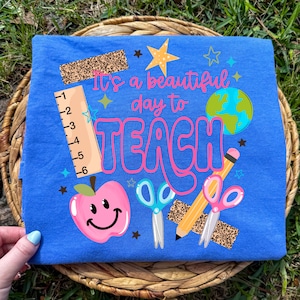 May include: A blue t-shirt featuring a graphic with the text "It's a beautiful day to TEACH" in pink. The design includes a ruler marked in inches, a pink smiling apple, a blue pair of scissors, a yellow pencil, a globe, and leopard print accents.