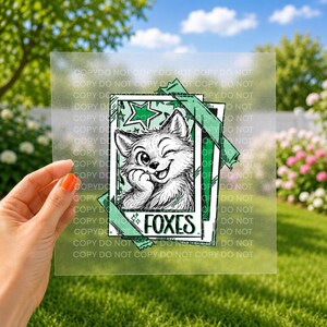 May include: A square, semi-transparent transfer sheet featuring a black and white line art illustration of a winking fox cub inside a rectangular frame. The design includes green accents, a star, and the word FOXES printed in a bold, black font.