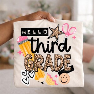 May include: A cream-colored t-shirt featuring a graphic with the text HELLO third GRADE. The design includes a yellow pencil, a pink pair of scissors, a leopard print star, leopard print lettering, black hearts, and a smiling face icon.