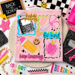 May include: A light pink t-shirt featuring colorful school-themed graphics including a composition notebook, pink scissors, a yellow pencil, a pushpin, and a pink apple. The text reads Radiate positivity, Be kind, and keep learning.