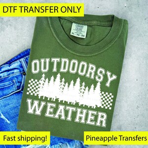 May include: An olive green t-shirt with the words "OUTDOORSY WEATHER" in white block letters, with a forest graphic. The shirt is folded on top of blue denim shorts. The tag reads XL-TG-EG.