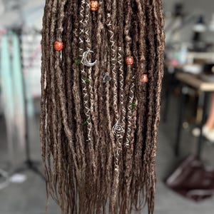 Puede incluir: Un conjunto de rastas sintéticas marrones con una apariencia texturizada. Las extensiones de cabello están adornadas con dijes plateados, incluyendo una luna creciente y formas de espiral, junto con pequeñas cuentas de madera e hilos blancos.