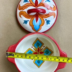 May include: A white ceramic bowl with red handles and a colorful geometric design. The bowl is 8 inches in diameter.