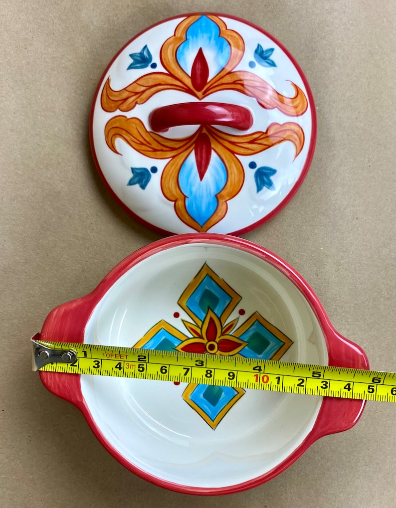 May include: A white ceramic bowl with red handles and a colorful geometric design. The bowl is 8 inches in diameter.