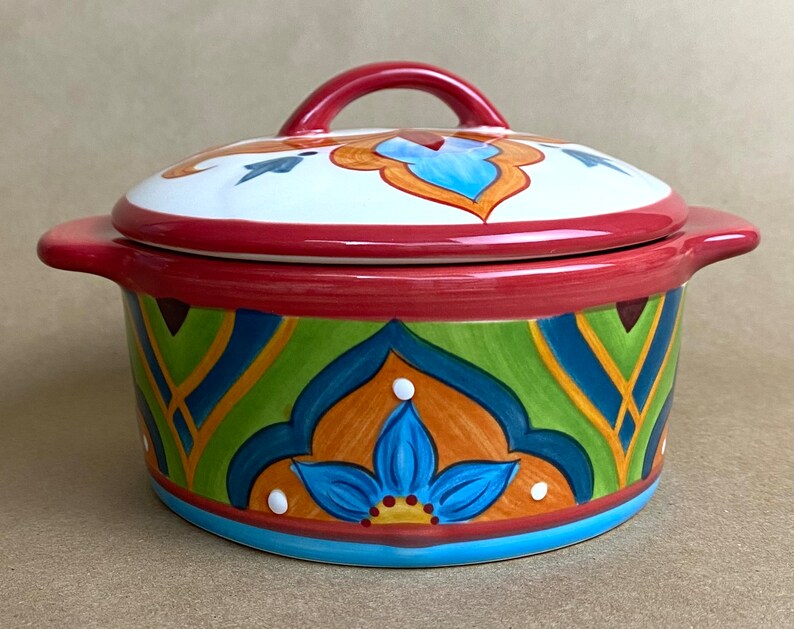 May include: A colorful ceramic casserole dish with a lid. The dish has a red rim and a blue, green, orange, and yellow geometric pattern. The lid has a red handle and a white center with a blue and orange design.