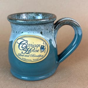 May include: A ceramic mug with a teal and white drip glaze finish. The front features an oval gold medallion with the text Carriage House Bed and Breakfast Jefferson, Texas and a small illustration of a carriage with a lantern.