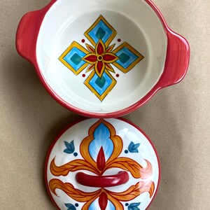 May include: A red ceramic casserole dish with a lid. The dish has a white interior with a blue, yellow, and red geometric design. The lid has a similar design with a red handle.