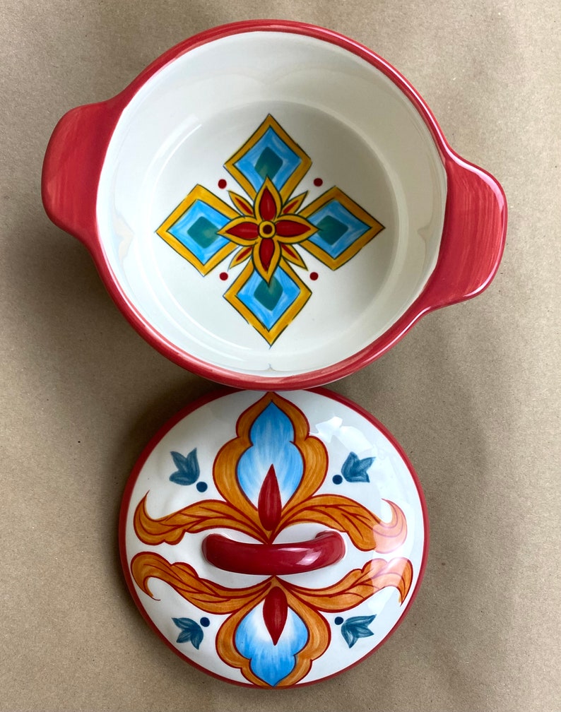 May include: A red ceramic casserole dish with a lid. The dish has a white interior with a blue, yellow, and red geometric design. The lid has a similar design with a red handle.