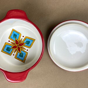 May include: A red ceramic baking dish with a white interior and a colorful geometric design. The dish has two handles and a matching lid.