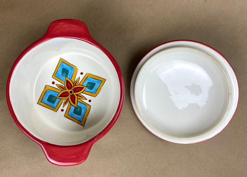 May include: A red ceramic baking dish with a white interior and a colorful geometric design. The dish has two handles and a matching lid.