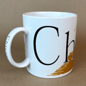May include: White ceramic mug with a large black "Ch" and a gold-colored illustration. The handle has "Chicago" printed on it. The mug is a standard size and shape, suitable for coffee or tea.