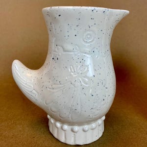May include: White ceramic jug with a speckled glaze and a floral design. The jug has a unique handle shaped like a bird's wing.