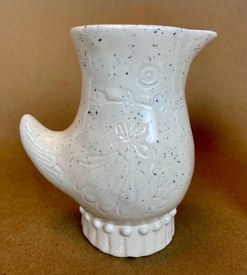 May include: White ceramic jug with a speckled glaze and a floral design. The jug has a unique handle shaped like a bird's wing.