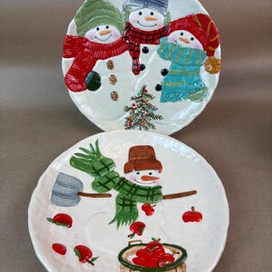 May include: Two white ceramic plates with snowman designs. One plate features three snowmen with hats and scarves, and a small Christmas tree. The other plate has a snowman with a green scarf, brown hat, and apples.
