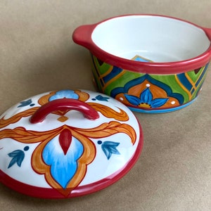 May include: A white ceramic bowl with a red rim and a colorful floral design. The bowl has a matching lid with a red handle.